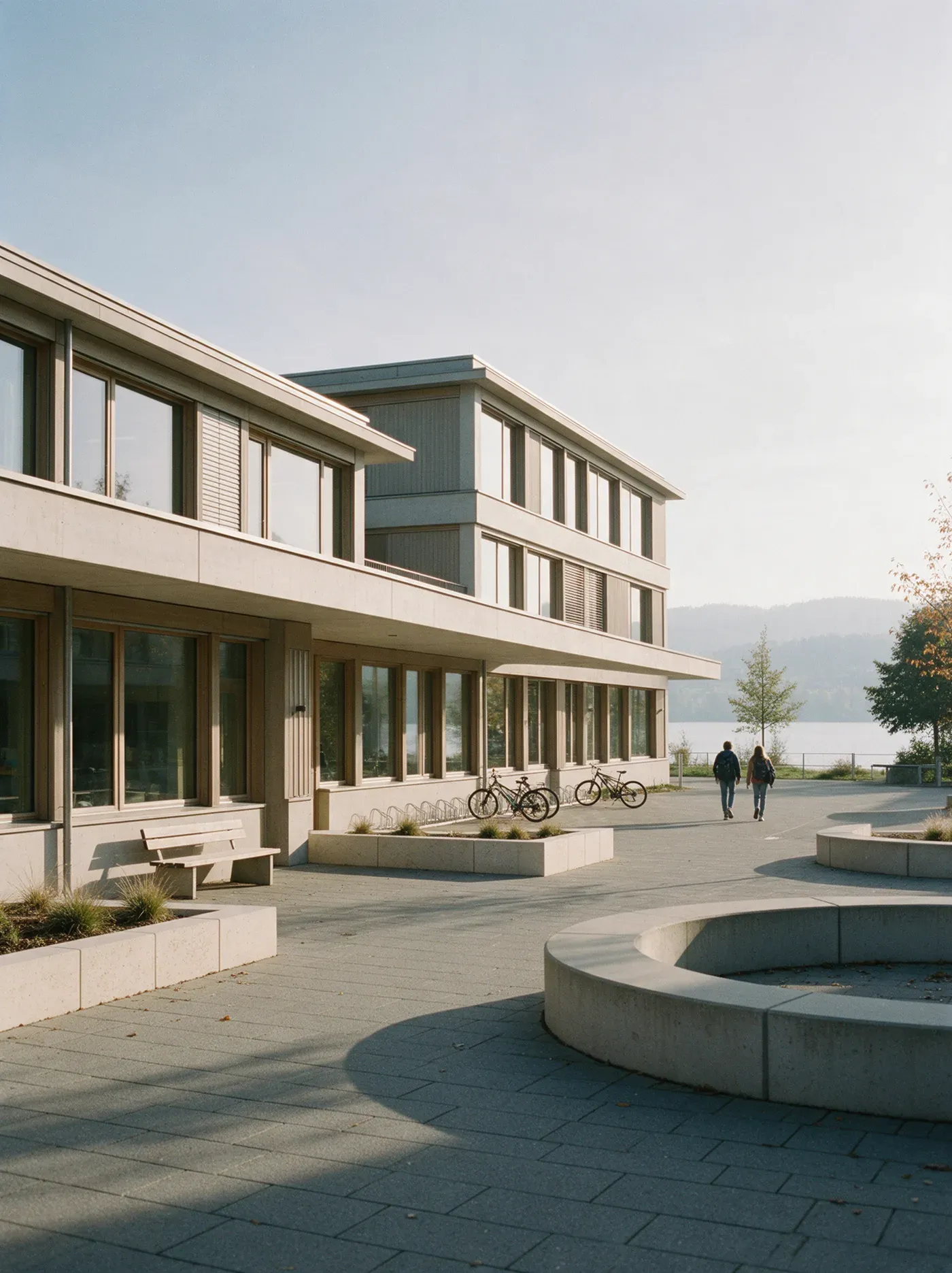 Ein helles Schulhaus mit Pausenplatz an einem sonnigen Herbstmorgen, im Hintergrund der See.